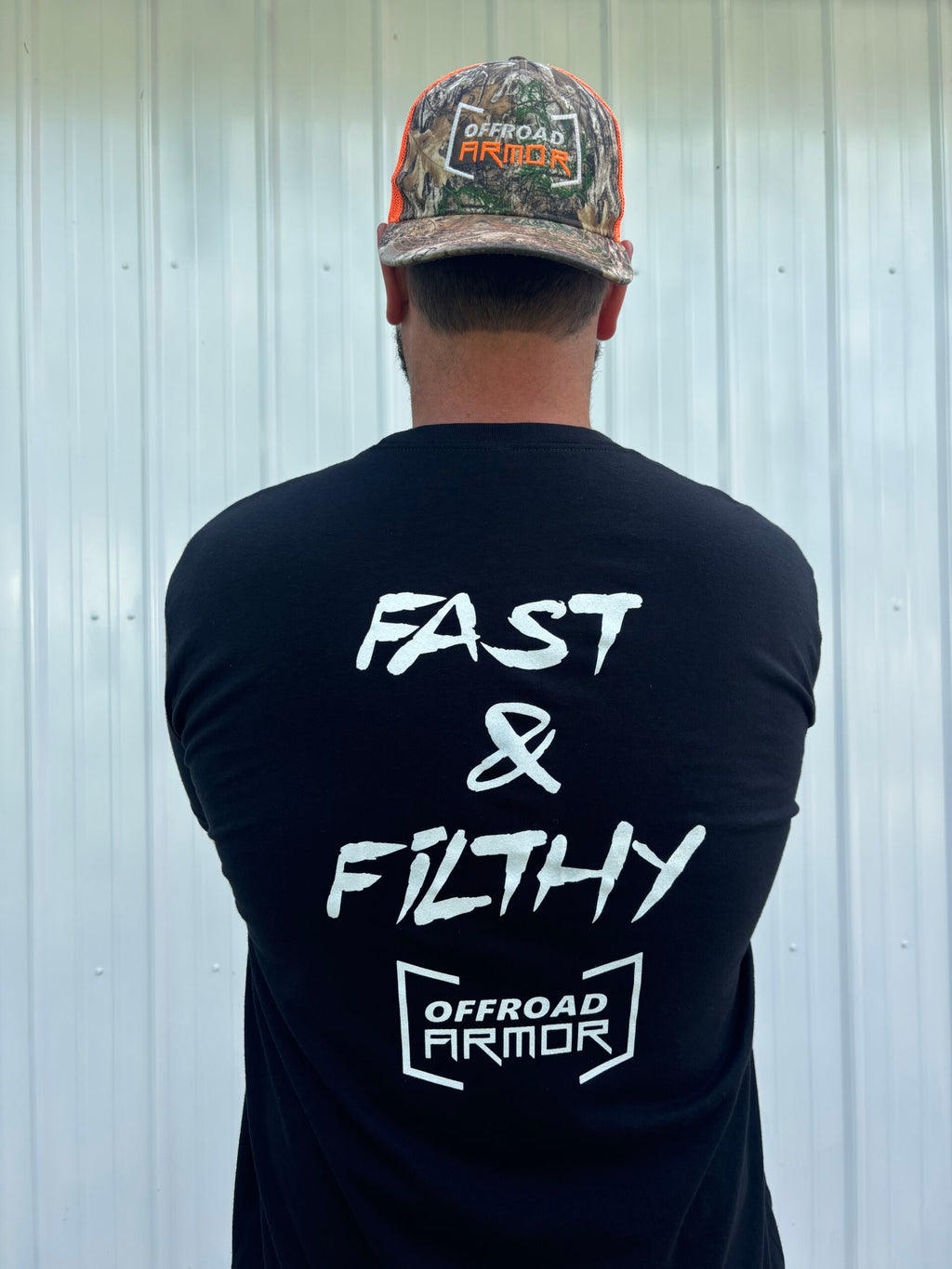 A bearded person faces away, wearing a Fast and Filthy - Offroad Armor Next Level T-Shirt with bold white “FAST & FILTHY” and “OFFROAD ARMOR” text, plus a camo hat featuring the OFFROAD ARMOR logo, against a white metal wall.