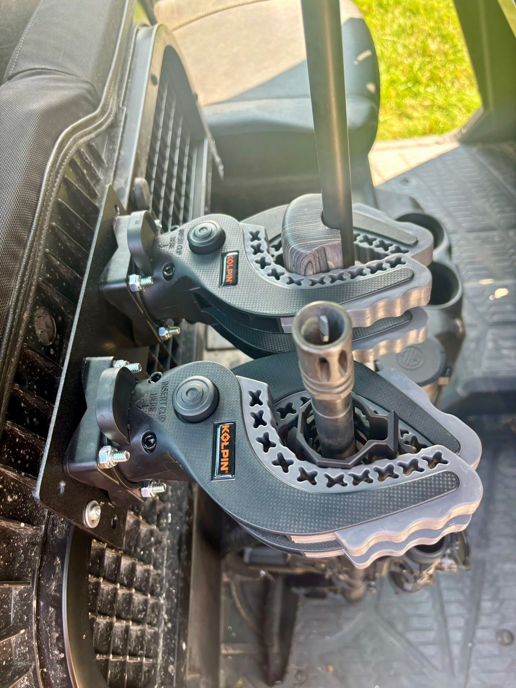 Close-up of an Offroad Armor Polaris Ranger Under Seat Hidden Rifle Mount securing a firearm inside a vehicle. The gray and black clamps attach to a rack, with the floor mat and seat visible in the background.