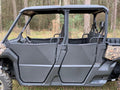 A side view of Can-Am Defender HD5/HD7/HD8/HD9/HD10 Max Half Doors in black with camouflage accents, installed on a four-door UTV parked on grass. The doors are closed, showing a spacious two-row cabin with black seats and trees in the background.