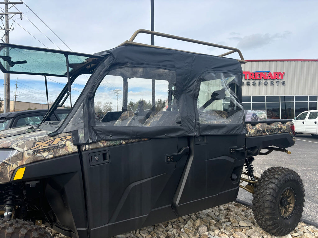 A Polaris Ranger Crew 900/1000 (2014+) with soft upper doors is parked on rocks outside a Trenary Resorts building. The UTV features black doors with clear plastic windows, while other vehicles and a cloudy sky are in the background.