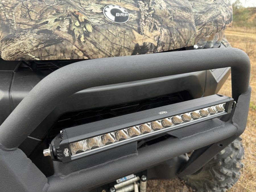 Close-up of a Can Am Defender 16″ Bumper Mount LED Light Bar Kit (not for Lonestar edition), installed on an off-road vehicle with a camouflage hood, BRP logo, sturdy black bumper, visible winch, tire, and grassy outdoor backdrop.