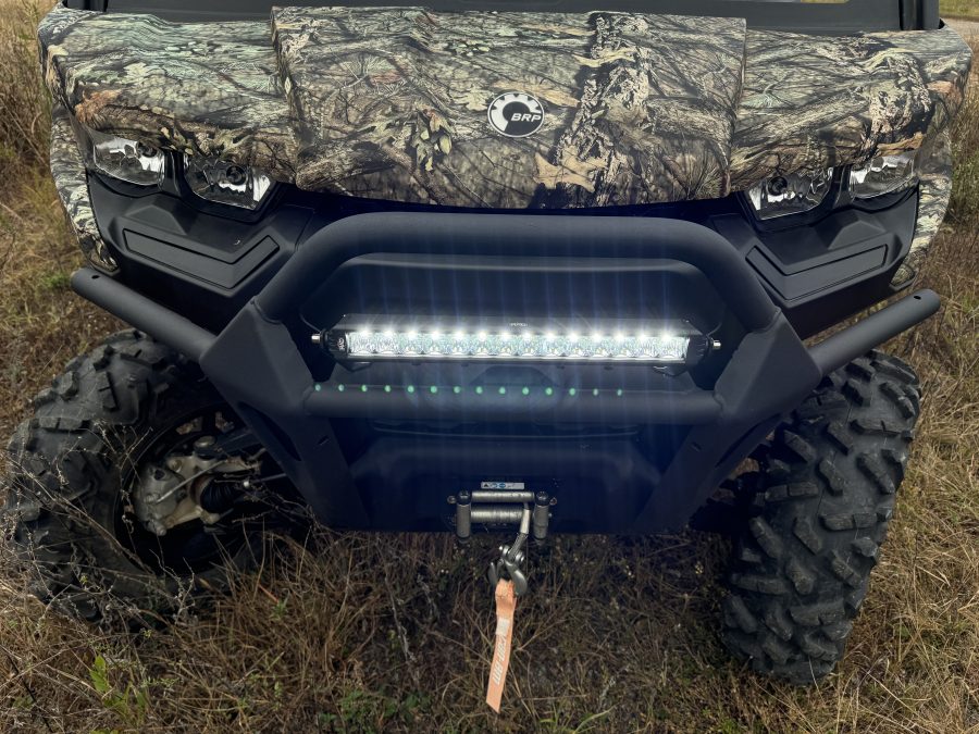 Close-up of a Can Am Defender with a 16″ Bumper Mount LED Light Bar Kit (not for Lonestar edition), camouflage paint, rugged tires, sturdy bumper, winch with orange strap, and BRP logo on the hood, parked on grass.