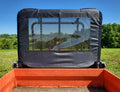 View from the bed of a utility vehicle with a Kubota RTV 900/1120 - Soft Back Panel, featuring a mesh and fabric rear window. Grassy fields and green trees are visible through it under a clear blue sky on a sunny day.