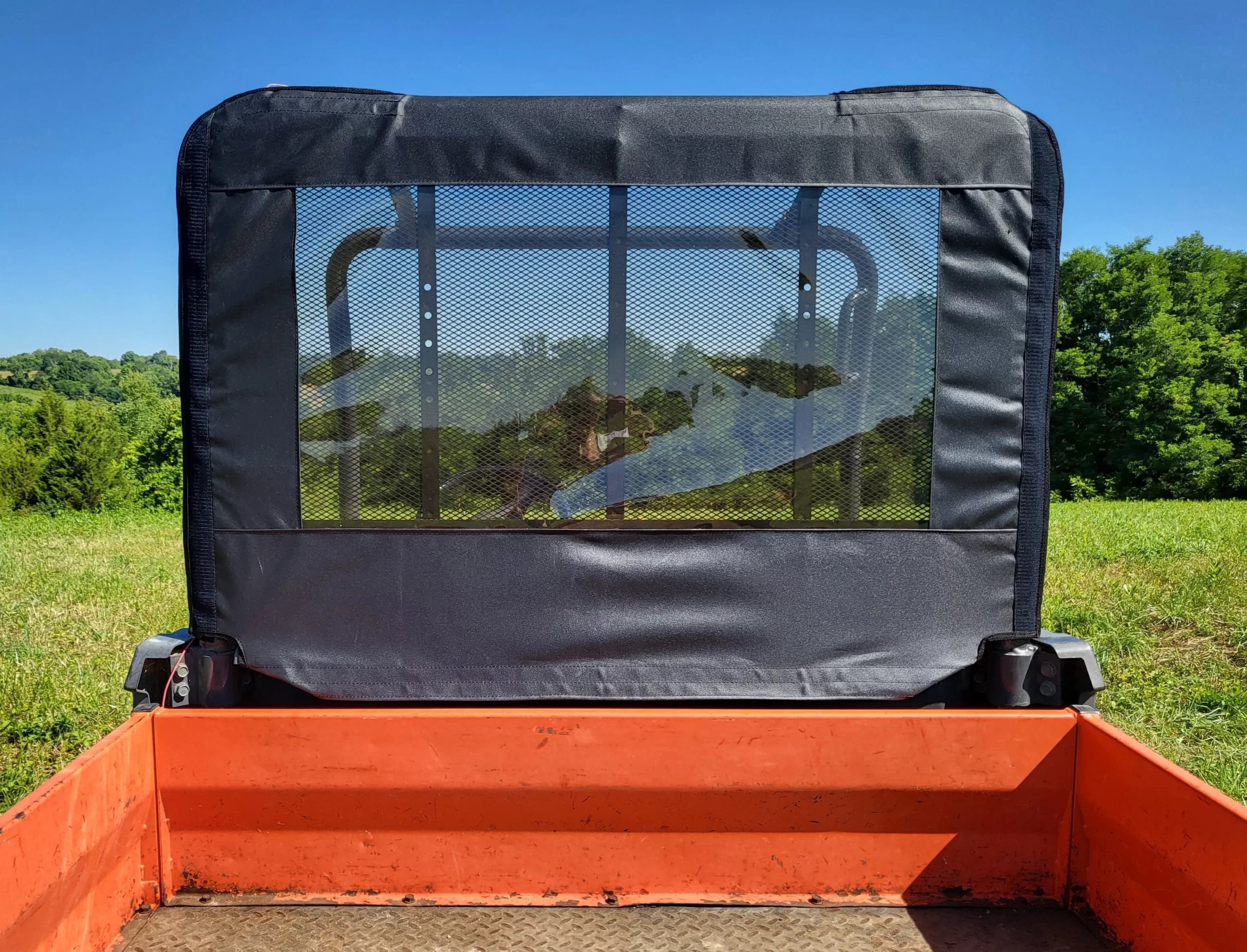View from the bed of a utility vehicle with a Kubota RTV 900/1120 - Soft Back Panel, featuring a mesh and fabric rear window. Grassy fields and green trees are visible through it under a clear blue sky on a sunny day.