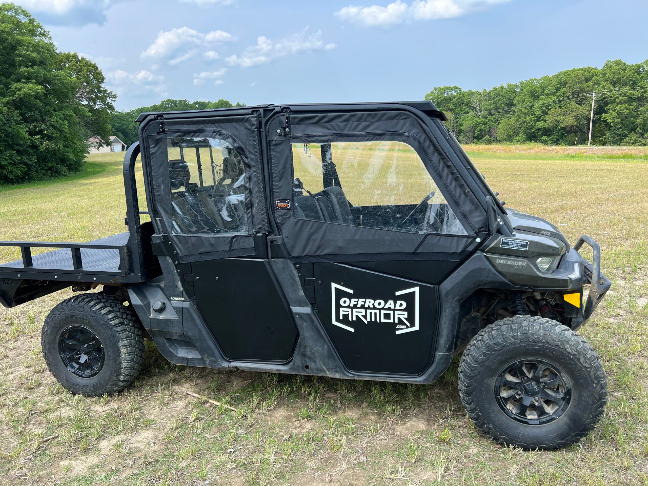 A Can-Am Defender Max with Framed Upper Soft Doors for ThumperFab Lower doors is parked on grassy terrain. It features clear plastic windows, rugged tires, and a metal cargo bed, with green trees and a cloudy blue sky in the background.