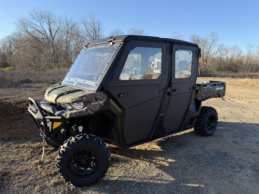 A rugged Can Am Defender UTV with four doors and off-road tires, fitted with a 16″ Bumper Mount LED Light Bar Kit (not for Lonestar edition), is parked on gravel amid bare trees and dry grass, evoking a sunny late fall or winter rural setting.