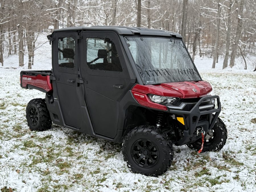 A Can Am Defender equipped with a 16″ Bumper Mount LED Light Bar Kit (not for Lonestar edition) is parked on snowy grass in a leafless forest, featuring large rugged tires, a front bumper guard, and a covered cab with clear windows.