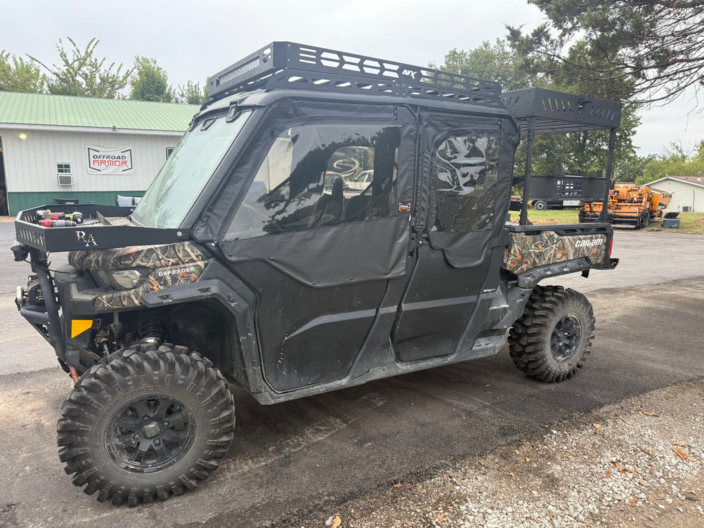 A Can-Am Defender Max with OEM half doors and Framed Upper Soft Doors is parked on pavement. It features rugged tires, camo accents, roof and cargo racks, with trees and a green metal building in the background.
