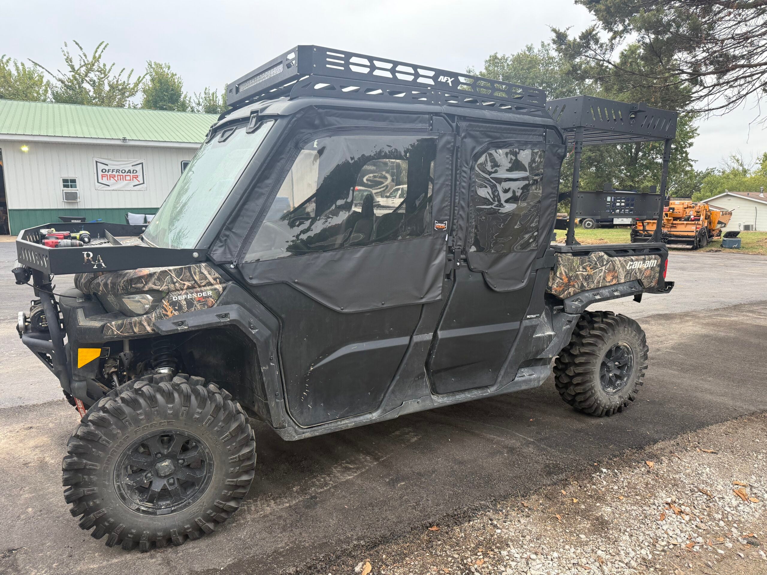 A Can-Am Defender Max with OEM half doors and Framed Upper Soft Doors is parked on pavement. It features rugged tires, camo accents, roof and cargo racks, with trees and a green metal building in the background.