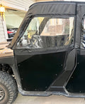 A close-up side view of a Polaris Ranger 900/1000 (2014+) fitted with the Soft Upper Door Kit, featuring a black zippered door with a clear plastic window. The vehicle is parked in a driveway near a house, with another car and garage in the background.