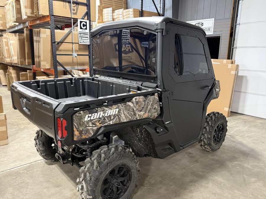 A black Can-Am UTV equipped with the Defender Full Aluminum Door Kit (converts from full to half, 2 door) is parked indoors on a concrete floor, with rugged tires and a large cargo bed. Shelves and boxes are visible in the background.
