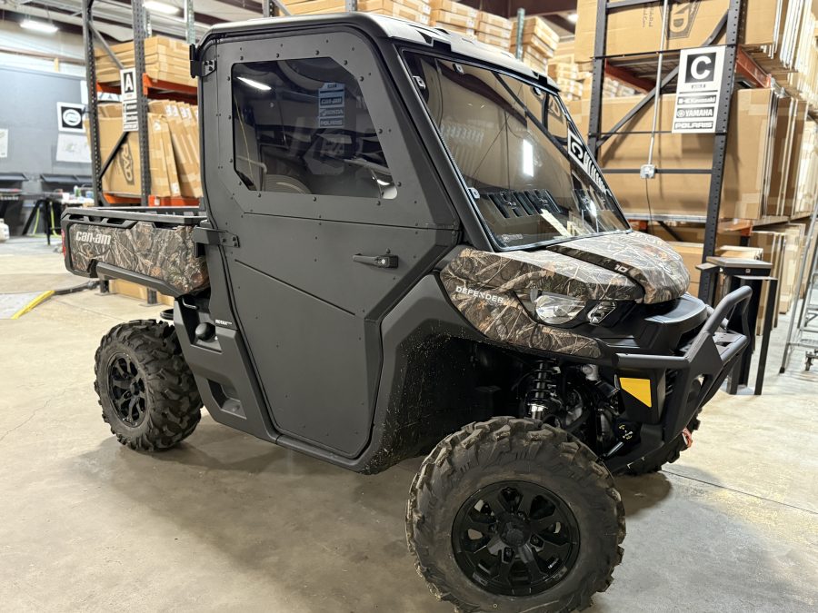 A Can-Am Defender with full aluminum doors (convertible to half), black with camo accents and large off-road tires, is parked indoors on concrete near shelves of brown boxes. The closed cab features tinted windows and “Can-Am” branding.