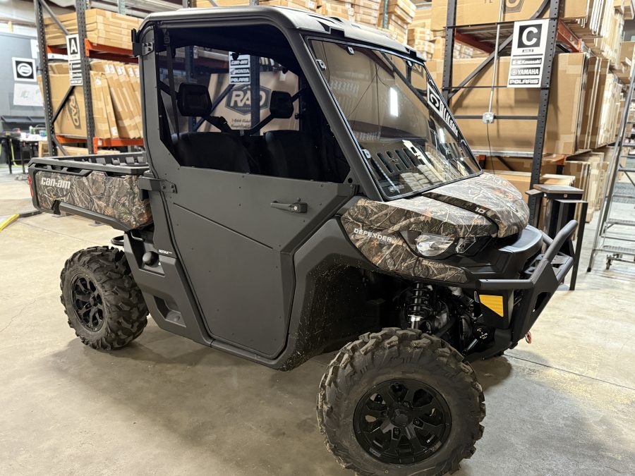 A Can-Am Defender side-by-side, equipped with the Can Am Defender Full Aluminum Door Kit (converts from full to half, 2 door), is parked indoors on concrete. Large off-road tires, a black cab, and cargo bed are visible amid warehouse shelving and boxes.