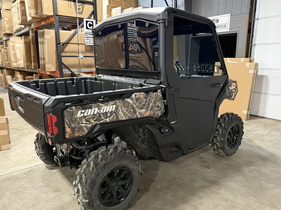 A Can Am Defender fitted with the Full Aluminum Door Kit (convertible from full to half doors) is parked indoors in a warehouse with shelves of boxes; the rear window is open, showing various cab controls.
