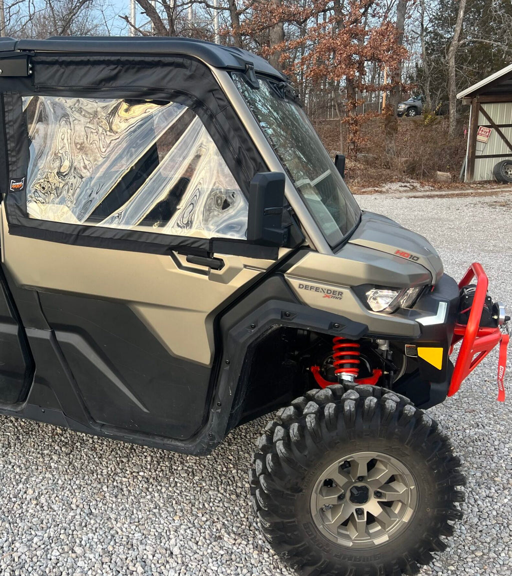 A Can-Am Defender with 2023+ Newer Style OEM Soft Framed Upper Doors sits on a gravel lot, featuring camo green color, black doors, clear windows, red suspension springs, and a red front bumper. Trees and a shed can be seen in the background.