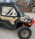 A Can-Am Defender with 2023+ Newer Style OEM Soft Framed Upper Doors sits on a gravel lot, featuring camo green color, black doors, clear windows, red suspension springs, and a red front bumper. Trees and a shed can be seen in the background.