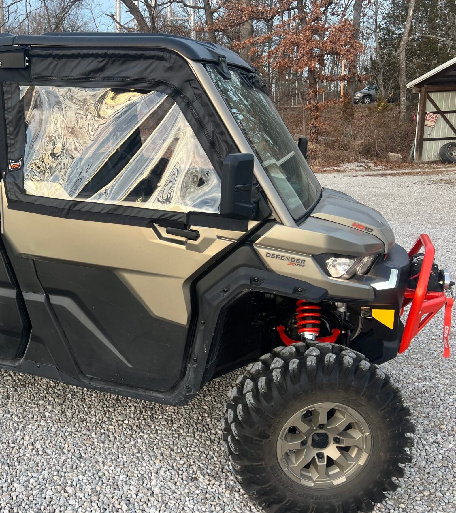 A Can-Am Defender with 2023+ Newer Style OEM Soft Framed Upper Doors sits on a gravel lot, featuring camo green color, black doors, clear windows, red suspension springs, and a red front bumper. Trees and a shed can be seen in the background.