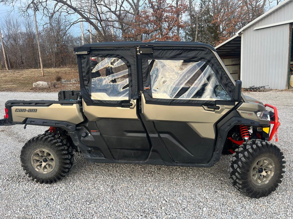 A side view of a Can-Am Defender Max with Framed Upper Soft Doors for OEM half doors, featuring a tan and black closed cab with plastic windows, parked on gravel by a metal building with leafless trees and brown grass in the background.