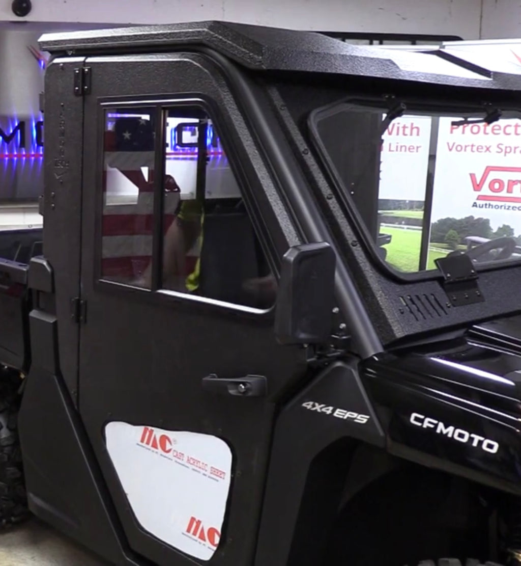 A black CF Moto Uforce 1000 with Armor Tech Full Steel Doors is parked indoors. The hard-sided doors have window inserts, through which part of an American flag and commercial signage are visible inside the building.