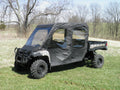 A John Deere Gator Xuv 4 Seater with a soft full cab enclosure and vinyl windshield, equipped with off-road tires, is parked on grass amid leafless trees and rolling green fields beneath a partly cloudy sky.