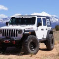 A white 2018+ Jeep JL Rubicon with Stealth Front Winch Bumper (Mid Width, Texture Black, Warn Zeon) is parked on dirt. Snow-capped mountains and a blue sky are in the background, with ROAD ARMOR on the windshield and gear mounted up front.