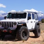 A white 2018+ Jeep JL Rubicon with Stealth Front Winch Bumper (Mid Width, Texture Black, Warn Zeon) is parked on dirt. Snow-capped mountains and a blue sky are in the background, with ROAD ARMOR on the windshield and gear mounted up front.