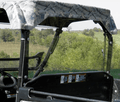 An Arctic Cat Prowler with a camouflage roof and no doors is shown sideways, featuring a Lexan Back Panel, clear windshield, and side panels. Green trees, grassy fields, and a clear sky form the background.