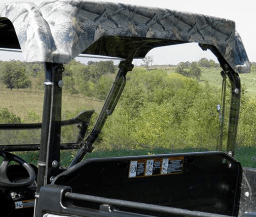 An Arctic Cat Prowler with a camouflage roof and no doors is shown sideways, featuring a Lexan Back Panel, clear windshield, and side panels. Green trees, grassy fields, and a clear sky form the background.