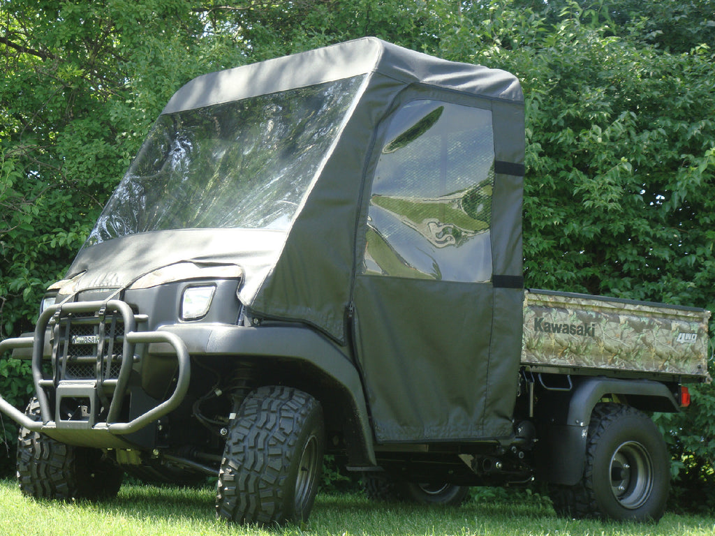 A Kawasaki Mule 3000/3010/4000/4010 with a Soft Full Enclosure and Vinyl Windshield sits on grass, featuring large tires and canvas sides, with dense green trees and bushes in the background.