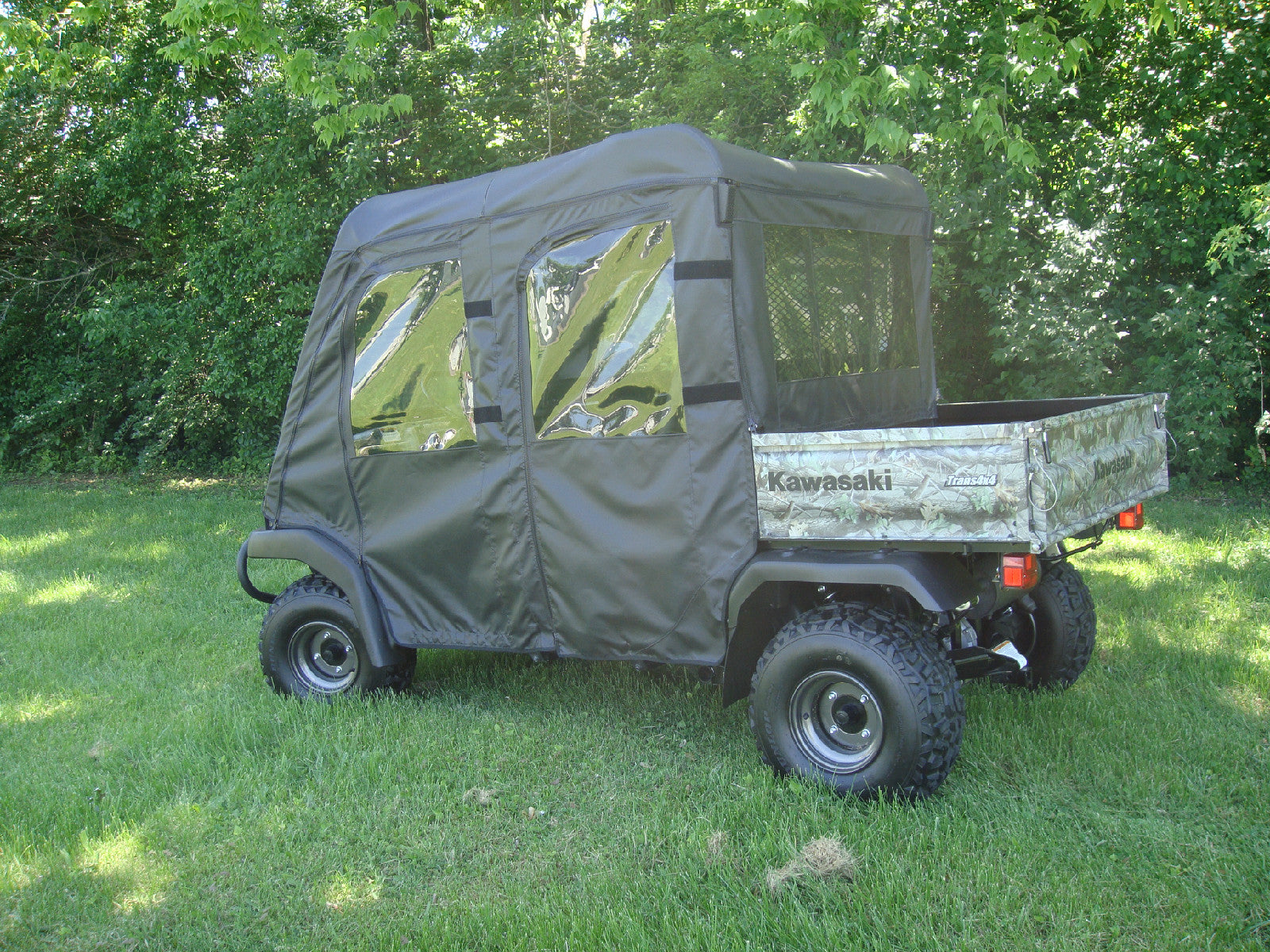 A green Kawasaki utility vehicle with large rugged tires and a camouflage cargo bed is parked on grass among trees, featuring the Mule Trans Soft Doors, Rear Window and Top Combo for an existing hard windshield covering the cab.
