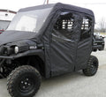 A black Kawasaki Mule Pro-Fxt fitted with a Soft Full Cab Enclosure with Vinyl Windshield and large off-road tires is parked on a gravel lot near industrial buildings and several other vehicles.