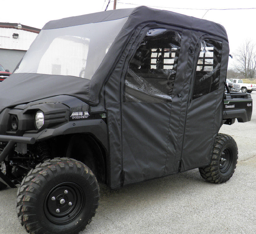 A black Kawasaki Mule Pro-Fxt fitted with a Soft Full Cab Enclosure with Vinyl Windshield and large off-road tires is parked on a gravel lot near industrial buildings and several other vehicles.