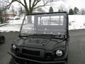 A black Kawasaki UTV with a Mule Pro-Fxt Split Lexan Windshield is parked on wet asphalt, light snow covering the ground and trees under an overcast sky, and buildings partially visible in the background.