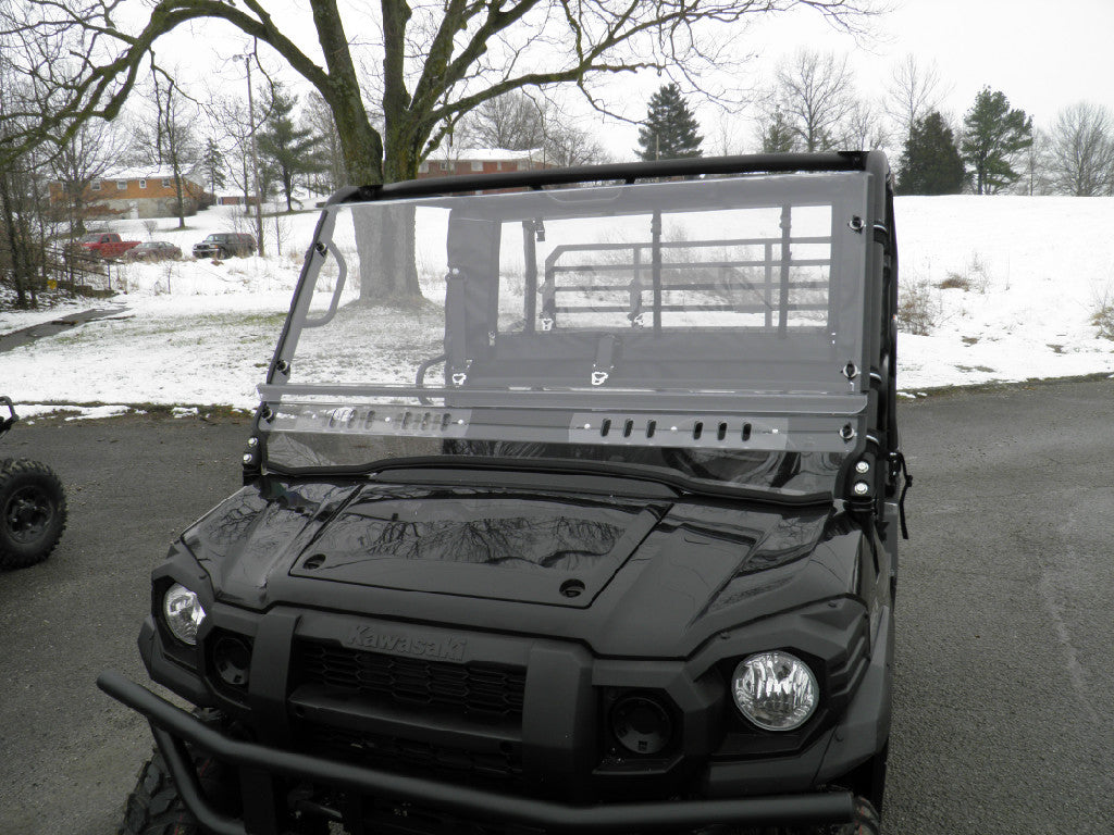 A black Kawasaki UTV with a Mule Pro-Fxt Split Lexan Windshield is parked on wet asphalt, light snow covering the ground and trees under an overcast sky, and buildings partially visible in the background.
