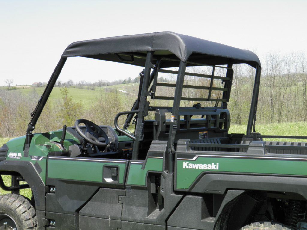 A Kawasaki Mule Pro-Fx Soft Top in green, featuring an open cab, black roof, and roll bars, is parked on grass with visible steering wheel and seats. Rolling hills, trees, and a clear sky provide a rural backdrop.