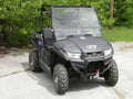 A black UTV featuring a Kymco 450I 2-Piece Vented Windshield (scratch resistant) with off-road tires, winch on the front bumper, and roll cage is parked on gravel, with trees and greenery visible in the background.