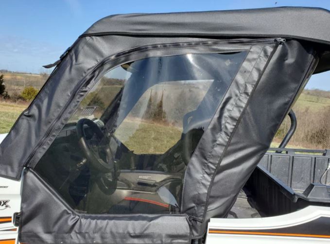 A close-up of a Kawasaki Teryx 800 (2-Seater) with black Soft Doors (Open Box Sale), featuring zippered enclosures and clear plastic windows. The steering wheel and dashboard are visible, set against grassy fields and a blue sky.