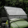 A Kawasaki Mule 600/610/SX Split Lexan Windshield with metal fasteners and center vents is mounted on a green utility vehicle with black trim, two gray seats, and dense green foliage in the background.