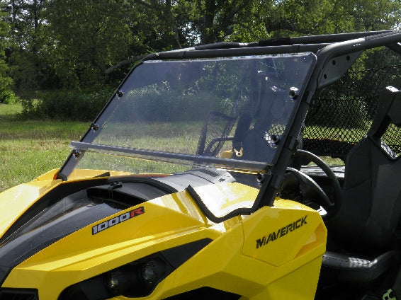 A Can-Am Maverick off-road utility vehicle with a Lexan windshield is parked on grass, featuring 1000R labels, black roll cage and seats. Trees and greenery appear in the background under daylight.