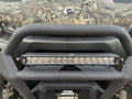 Close-up of a Can Am Defender 16″ Bumper Mount LED Light Bar Kit (not for Lonestar edition) installed on an off-road vehicle with a camouflage front, circular “BRP” logo, visible suspension, and undercarriage below.