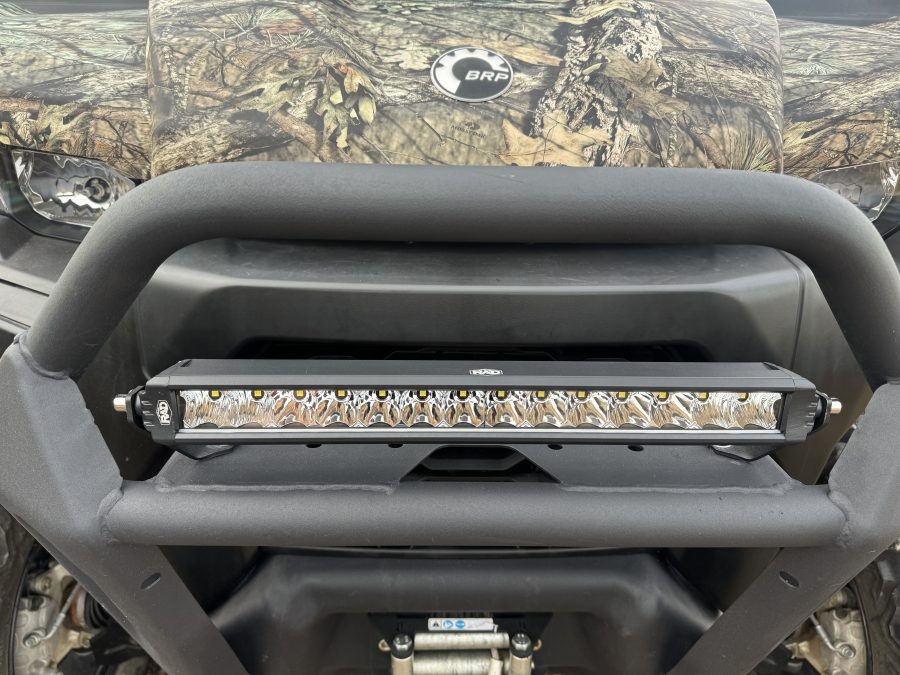 Close-up of a Can Am Defender 16″ Bumper Mount LED Light Bar Kit (not for Lonestar edition) installed on an off-road vehicle with a camouflage front, circular “BRP” logo, visible suspension, and undercarriage below.