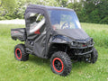 A Mahindra Mpact Xtv 750/1000 with large rugged tires and red accents is parked on grass. It features a Full Soft Enclosure With Vinyl Windshield, fully enclosing the cab. Trees and bushes are in the background under a partly cloudy sky.