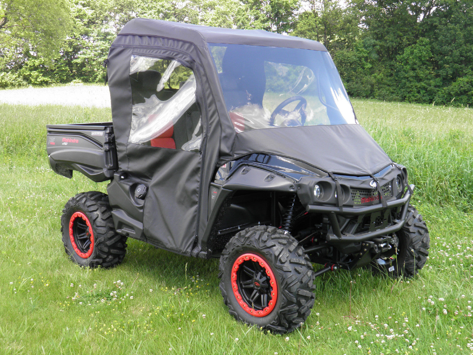 A Mahindra Mpact Xtv 750/1000 with large rugged tires and red accents is parked on grass. It features a Full Soft Enclosure With Vinyl Windshield, fully enclosing the cab. Trees and bushes are in the background under a partly cloudy sky.