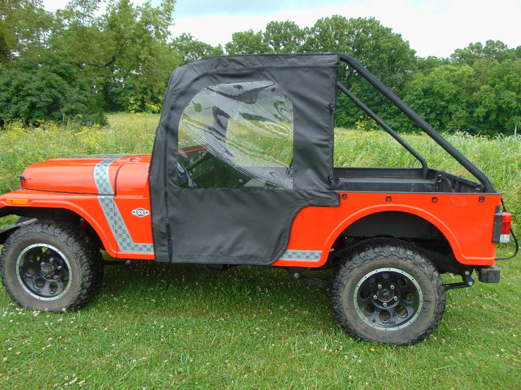 A Mahindra Roxor equipped with the Soft Door Kit featuring a black soft top and clear plastic window is parked on grass. Large tires, open cargo bed, and trees with wildflowers appear in the background under a cloudy sky.