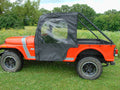 A Mahindra Roxor equipped with the Soft Door Kit featuring a black soft top and clear plastic window is parked on grass. Large tires, open cargo bed, and trees with wildflowers appear in the background under a cloudy sky.