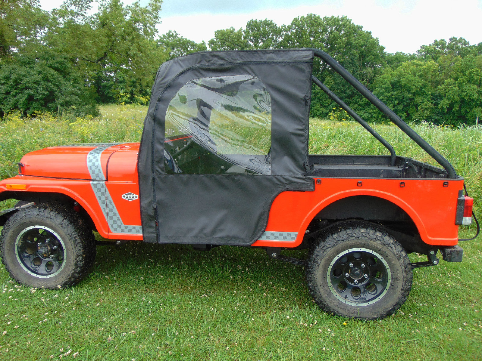 A Mahindra Roxor equipped with the Soft Door Kit featuring a black soft top and clear plastic window is parked on grass. Large tires, open cargo bed, and trees with wildflowers appear in the background under a cloudy sky.