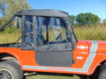 A close-up of a bright orange off-road Mahindra Roxor with “LIMITED EDITION” and “ROXOR” labels, featuring Upper Soft Doors with clear windows and a black soft-top, set against green grass, trees, and a blue sky.