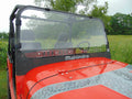 A close-up of a red Mahindra Roxor with a 1/4 Full Lexan Windshield shows water droplets on the windshield, two empty front seats, and the Mahindra logo on the dashboard. Trees and greenery blur in the background as it sits on grass.