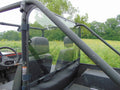 A close-up side view of a two-seater off-road utility vehicle with a Mahindra Roxor Lexan Rear Windshield, black seats, roll cage, and partial windshield, parked in a grassy field with trees under an overcast sky.