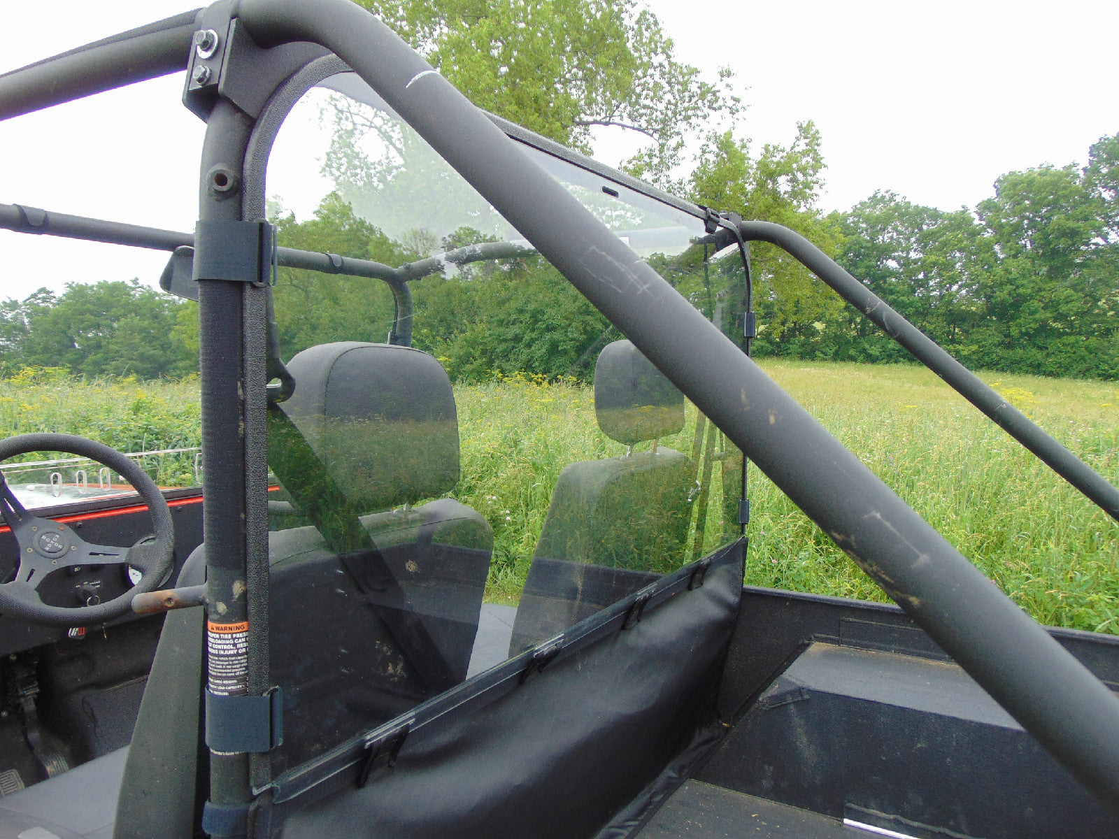 A close-up side view of a two-seater off-road utility vehicle with a Mahindra Roxor Lexan Rear Windshield, black seats, roll cage, and partial windshield, parked in a grassy field with trees under an overcast sky.